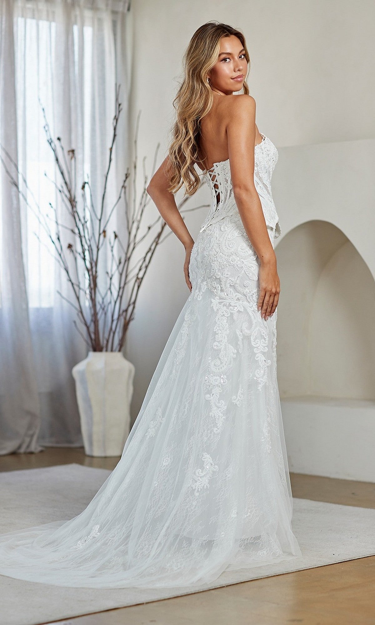 A woman wearing the Amelia Couture 2151 Long Prom Dress, featuring a strapless corset bodice with elegant lace embroidery, a fitted silhouette, and train, stands indoors looking over her shoulder.
